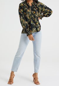 Černá květinová blůza s oranžovými a zelenými vzory, nařasený výstřih, nafouknuté dlouhé rukávy, spárovaná se světle modrými džíny a béžovými sandály na podpatku.