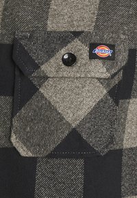 Plaid flannel shirt featuring gray and black colors, a buttoned chest pocket, and a Dickies brand label on the pocket. Textured fabric.