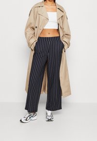 Beige trenchcoat, vit crop top, marinblå randiga vida byxor och silverfärgade sportskor med blå accenter, som har sidofickor och en avslappnad passform.