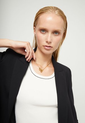 Femme blonde en blazer noir et haut blanc tenant un collier à chaîne dorée avec un pendentif abstrait sur un fond uni.
