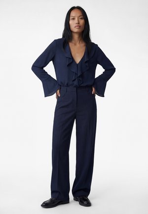 Femme debout les mains sur les hanches, portant un chemisier bleu marine à volants, un pantalon large à rayures bleu marine et des mocassins noirs sur un fond blanc.