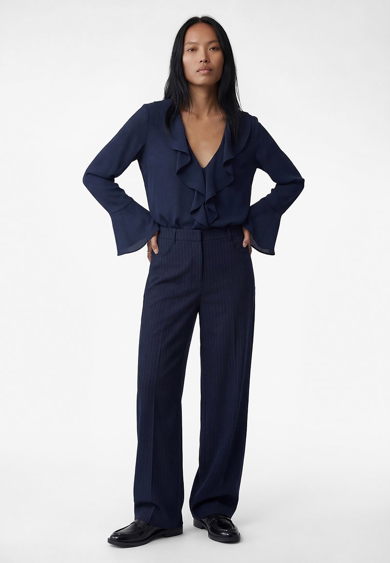 Kobieta stojąca z rękami na biodrach, ubrana w granatową bluzkę z falbankami, granatowe spodnie w szerokie prążki oraz czarne mokasyny na białym tle.