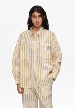 Kobieta ubrana w oversizową kremową koszulę i spodnie w pionowe brązowe paski, stojąca z jedną ręką w kieszeni na białym tle.