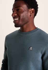 Dark teal sweatshirt with a round neckline, ribbed cuffs, and a small black and white logo on the chest. Smooth texture, casual fit.