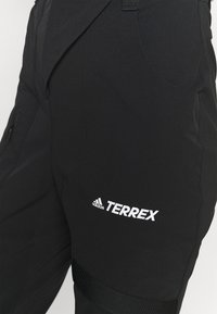 Pantalones exteriores negros con una textura suave y ligera. Cuentan con una cinturilla elástica y un logo blanco en el muslo. Incluyen bolsillos laterales.