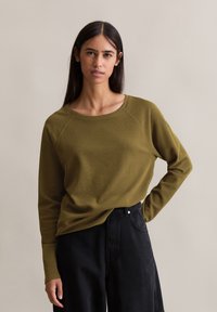 Oliven grønn langermet topp med rund hals og raglanermer, kombinert med høyt liv svarte bukser. Myk tekstur, uformell design.