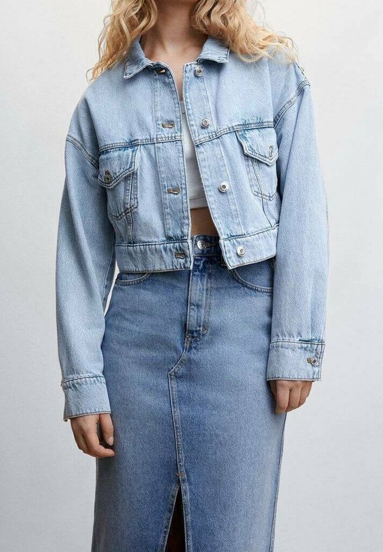Femme portant une veste courte en jean bleu clair sur un crop top blanc, associée à une jupe en jean bleu clair taille haute avec une fente à l'avant.