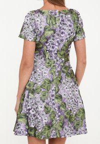Blommig klänning med korta ärmar, med lila och gröna blommönster på ett texturerat tyg. Utsvängd kjol med dragkedja i ryggen.
