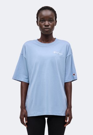 Kvinde iført en lys blå oversized Champion t-shirt med logo på brystet og ærmet, stående mod en ensfarvet lys baggrund.