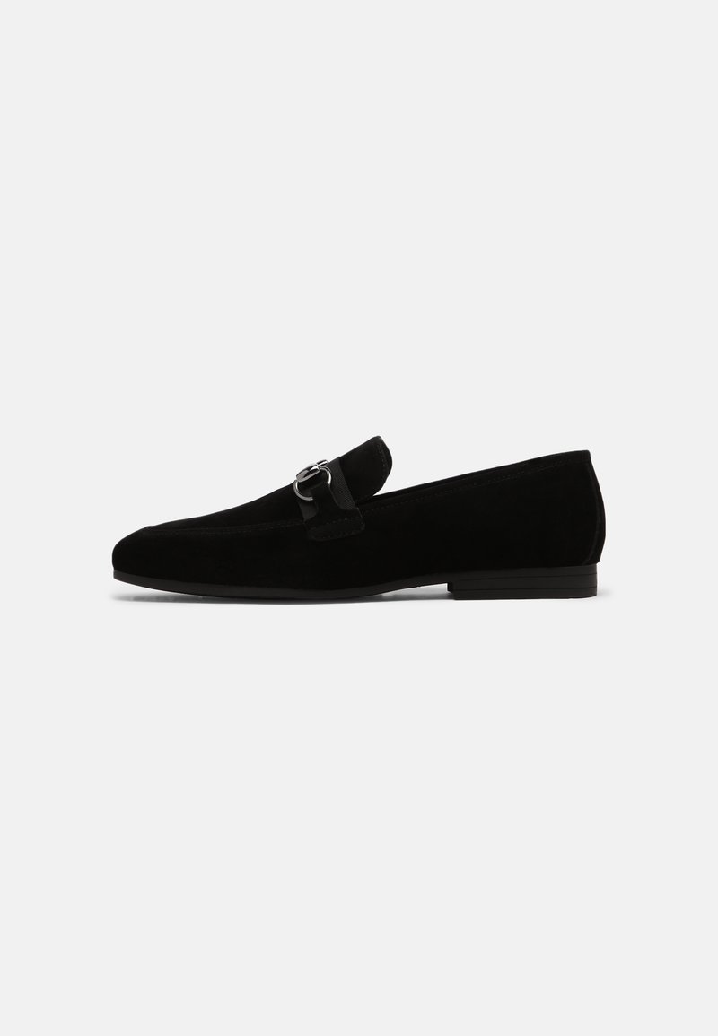 Mocassin homme en suédine noire avec détail mors argenté et petit talon superposé sur fond blanc uni.