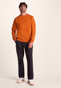 Man smiling, wearing an orange sweatshirt, dark pants, and white sneakers with black stripes, standing against a plain beige background.