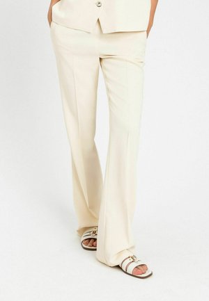 Parte inferiore del corpo di una persona che indossa pantaloni su misura color crema e sandali bianchi con fibbie dorate, in piedi davanti a uno sfondo bianco uniforme.