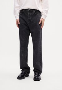 Person iført løse sorte jeans, sorte lædersko og en hvid langærmet bluse, der står mod en ensfarvet lys baggrund.