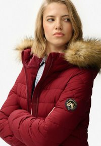 Chaqueta acolchada burdeos con capucha de pelo sintético, que cuenta con un cierre de cremallera y un parche circular con logo en la manga izquierda. Textura gruesa acolchada.