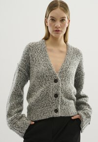 Grå stickad cardigan med v-ringning, långa ärmar och fem stora bruna knappar. Strukturerad tygkvalitet med lös passform och ribbad nederkant.