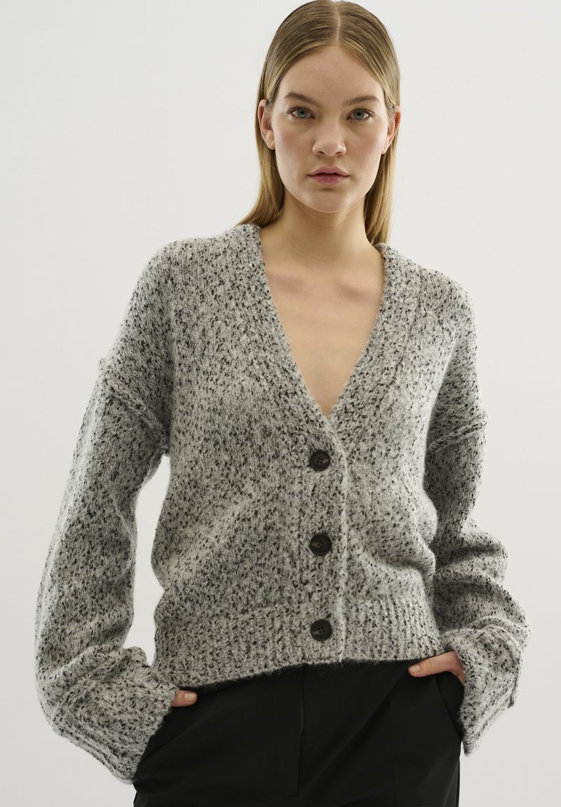 Grå stickad cardigan med v-ringning, långa ärmar och fem stora bruna knappar. Strukturerad tygkvalitet med lös passform och ribbad nederkant.