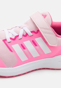Scarpa sportiva rosa con tomaia in rete, strisce bianche, un cinturino in velcro e suola texture. Gli accenti includono un contrasto di colori rosa e bianco.