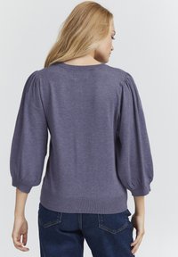 Lilla kampsärp, millel on puhvadega kolmveerand-sleeved varrukad ja soonikuline äär. Pehme kudumitekstuur. Kombineeritud tumesiniste teksadega, vaadatuna tagant.