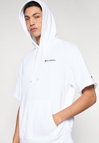 Λευκό φούτερ με κουκούλα που διαθέτει μπροστινή τσέπη, κοντά μανίκια και κορδόνια στη κουκούλα. Περιλαμβάνει ένα μικρό μαύρο κεντημένο λογότυπο στο στήθος.