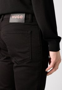 Jeans en denim noir avec une texture lisse, présentant une étiquette en cuir HUGO sur la ceinture et un design standard de poche arrière.