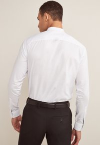 Hvid skjorte med lange ærmer og en mandarin-krave, kombineret med mørke bukser og et sort bælte, set bagfra.