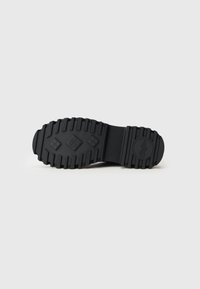 Suela de zapato de goma negra con surcos profundos y un patrón de relieve en forma de diamante, que presenta un logo circular cerca del talón.