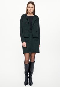 Zelený kostkovaný blazer s dvojitými knoflíky, zkombinovaný s odpovídající sukní a černými koženými vysokými botami.