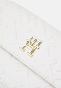 Hvid læderpung med et hævet geometrisk mønster og en guld logo-plakette, accentueret med detaljeret syning langs kanterne.