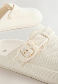 Witte klompen gemaakt van een gestructureerd materiaal met een gespband, afgeronde neus en een flexibele zool met een gelijnd patroon. Maat 12 aangegeven.