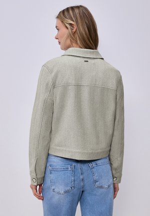 Femme aux cheveux blonds portant une veste gris clair à chevrons et un jean en denim bleu, tournée dos à l'appareil photo sur un fond uni.