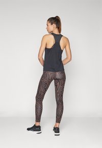 Débardeur de sport noir avec un dos nageur et une petite découpe à l'arrière, associé à un legging imprimé marron et des chaussures de sport noires.