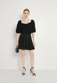 Mulher vestindo uma blusa preta texturizada com mangas balão, saia preta plissada, sapatos de salto pretos com tiras, segurando uma pequena bolsa preta, de pé contra uma parede simples.