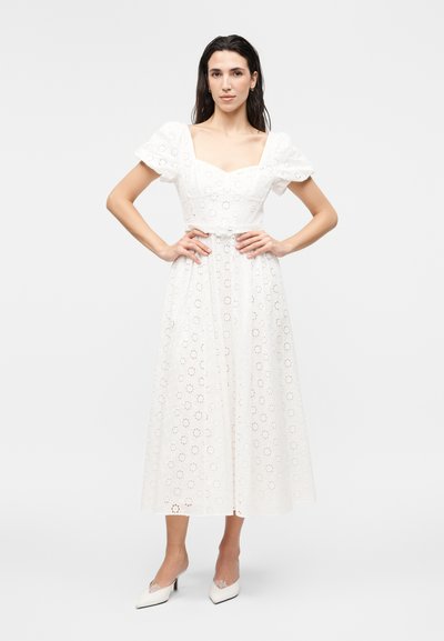 Γυναίκα στέκεται με τα χέρια στους γοφούς, φοράει λευκό μίντι φόρεμα με φουσκωτά μανίκια και φλοράλ δαντέλα με τρύπες, σε συνδυασμό με λευκές μυτερές γόβες.