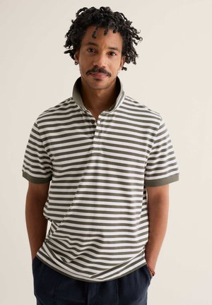 Mężczyzna z kręconymi włosami i wąsami, ubrany w zielono-białą pasiastą koszulkę polo, stoi z rękami w kieszeniach na jednolitym tle.