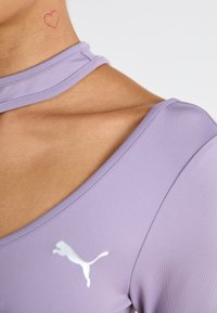 Lilla treningstopp med korte ermer, med en hvit Puma-logo på venstre bryst og et unikt halsdesign. Et lite rosa hjerte-tatovering synlig.