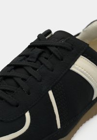 Close-up of a black sneaker with black laces and cream-colored stripes, showing textured fabric and detailed stitching.