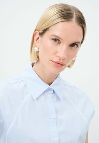 Camicia a righe blu chiaro con colletto e maniche corte, caratterizzata da una chiusura con bottoni e dettagli sottili sulle spalle. Tessuto morbido e liscio.