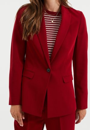 Femme portant un blazer rouge cintré avec un seul bouton noir sur un top rayé rouge et blanc, portant des colliers en or superposés.