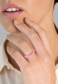 Rózsaszín zománcozott gyűrű aranyszínű kiemeléssel és egy kis drágakővel, ujjakon egy semleges háttér előtt.