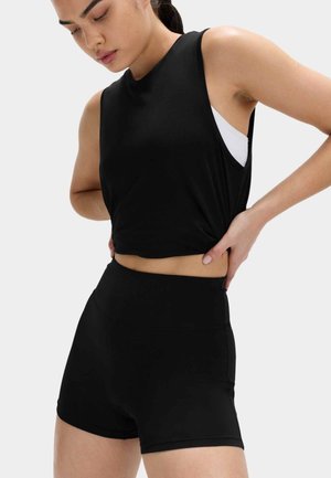 Kobieta poprawiająca czarny bezrękawnik crop top i czarne sportowe szorty z wysokim stanem, stojąca na jasnym jednolitym tle.
