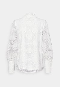 Vit spetsblus med blommönster, långa puffärmar och krage. Texturerad tyg med genomskinlig finish.