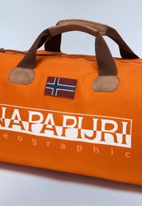 Oranssi duffel-laukku ruskeilla yksityiskohdilla, jossa on päällä sinivalkoinen lippu ja rohkeaa valkoista tekstiä "NAPAPIJRI geografia".