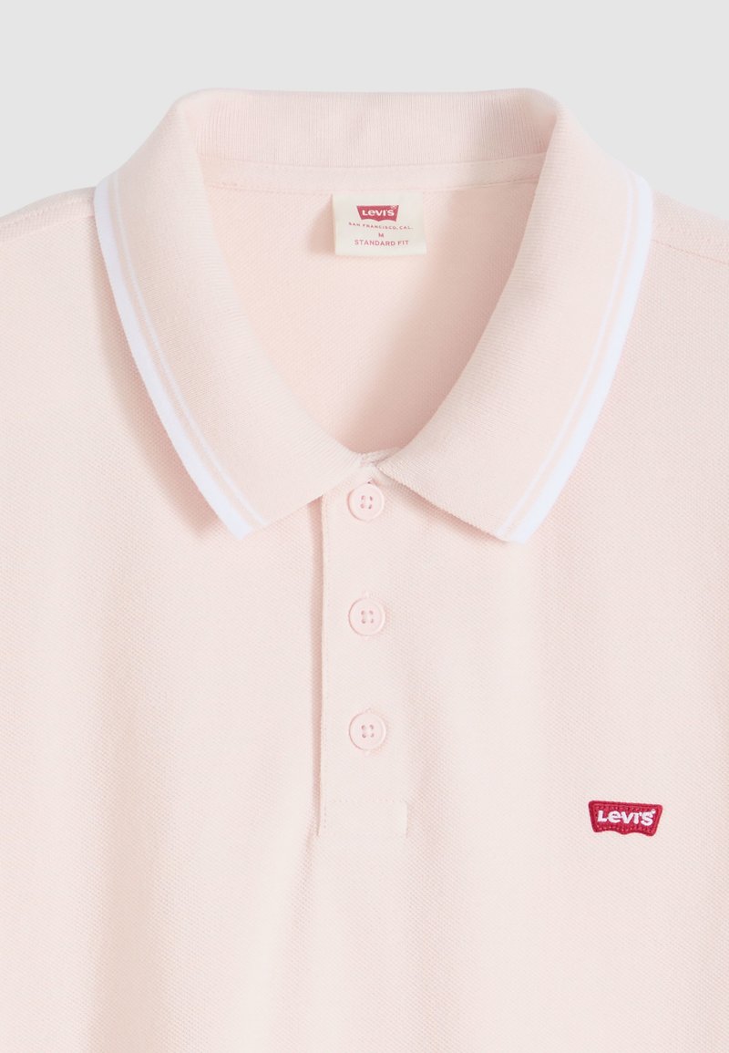 Polo Levi's rose clair avec bordure blanche au col, trois boutons sur le devant et logo rouge Levi’s sur la poitrine.