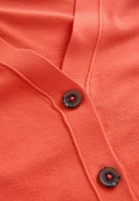 Pamučni kardigan koraljne boje s dizajnom V-izreza, s tamnim okruglim gumbom sprijeda i finom teksturom.