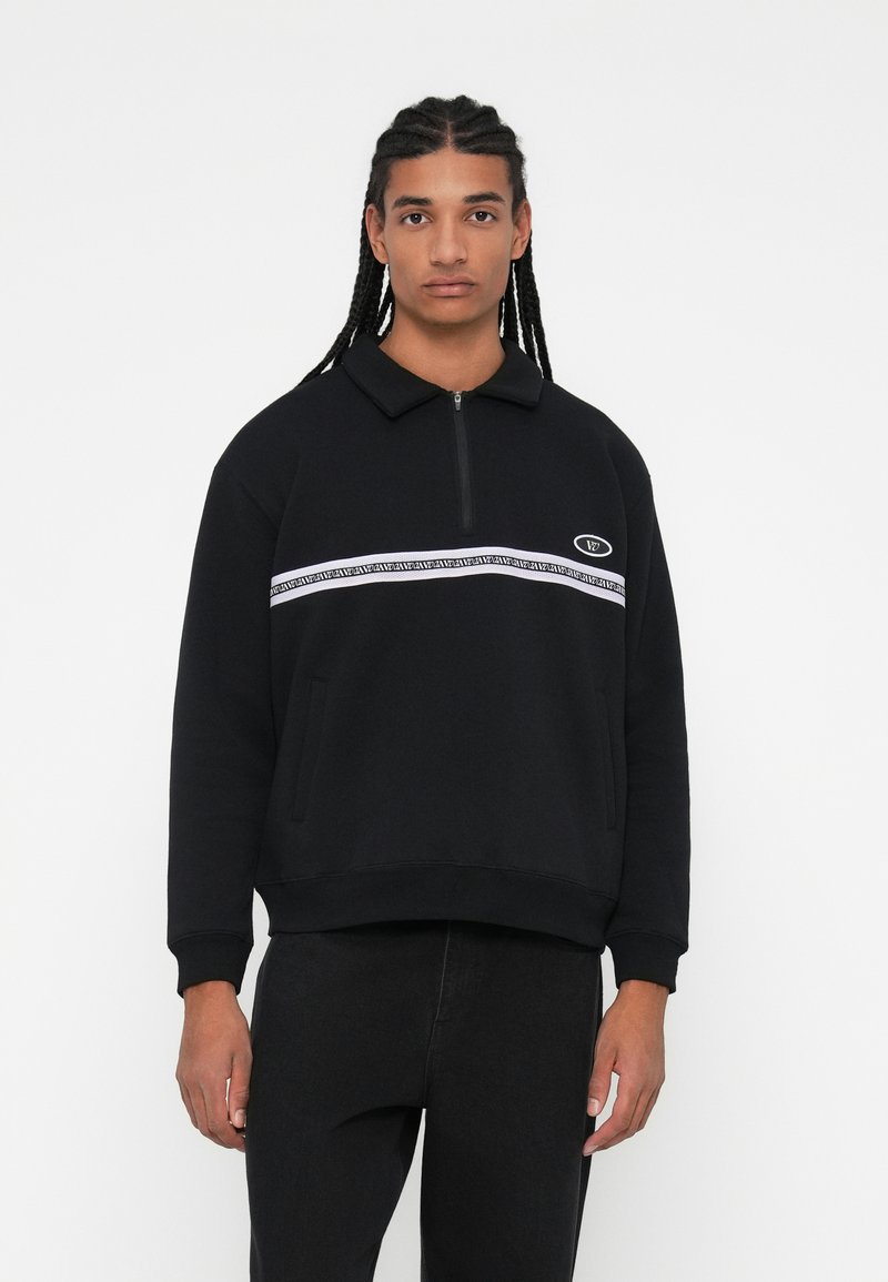 Sudadera negra con cuello y cremallera, con una franja horizontal blanca y logotipo, tejido texturizado y dos bolsillos laterales.