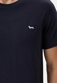 T-shirt in cotone blu navy con un piccolo logo ricamato di un bassotto bianco sul lato sinistro del petto. Maniche corte con scollatura a girocollo.
