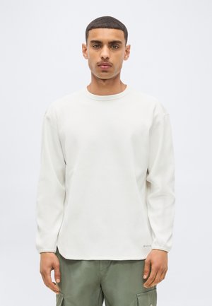 Giovane uomo con capelli corti che indossa una camicia bianca a maniche lunghe e pantaloni cargo verdi, in piedi davanti a uno sfondo chiaro e uniforme.