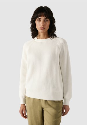 Kvinde med mørkt, bølget hår iført en hvid ribbet sweater og lysebrune bukser, stående foran en ensfarvet grå baggrund.