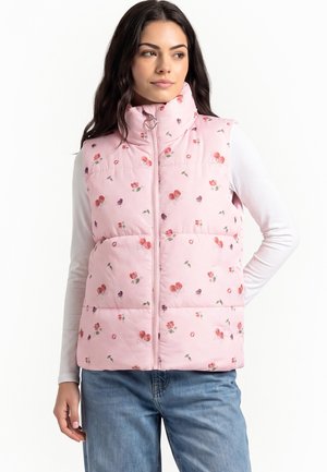 Vrouw die een roze gebloemd gewatteerd vest draagt met een rits-trekker in de vorm van een hartje, een witte lange mouw blouse en een blauwe spijkerbroek, staand met één hand achter de rug.