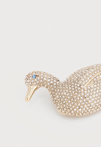 Παραμάνα σε σχήμα κύκνου από χρυσό, καλυμμένη με μικρά διαφανή κρυστάλλινα στοιχεία και έναν μοναδικό μπλε κρύσταλλο για το μάτι, τοποθετημένη πάνω σε λευκό φόντο.